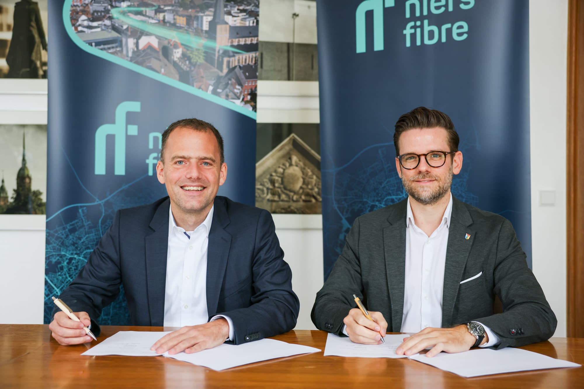 Zwei Männer sitzen an einem Tisch und unterschreiben Blätter, hinter ihnen stehen Niersfibre Plakate
